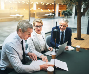 Three mature business executives discussing paperwork and using a laptop while having a meeting together in the lobby of an office building